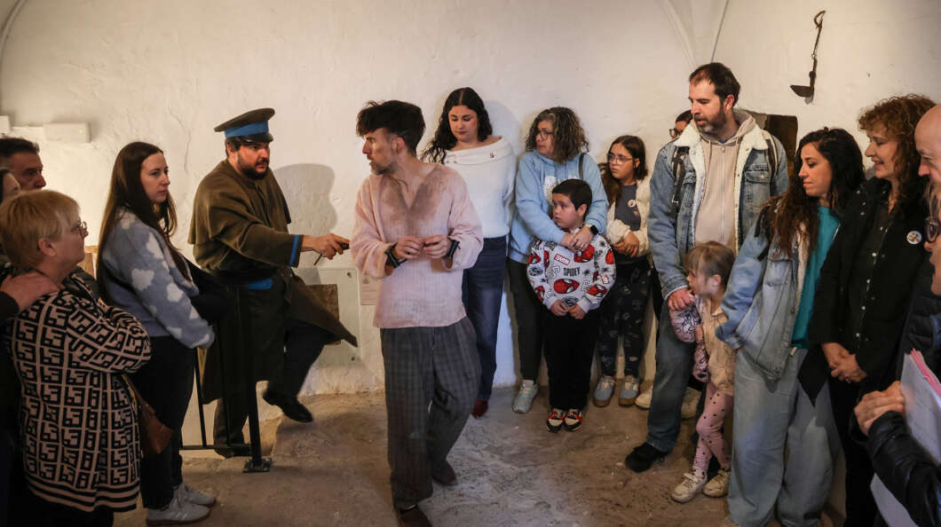 Group of adults and children watching a man in brown historical costume perform a gesture in a bare, white-walled room.
