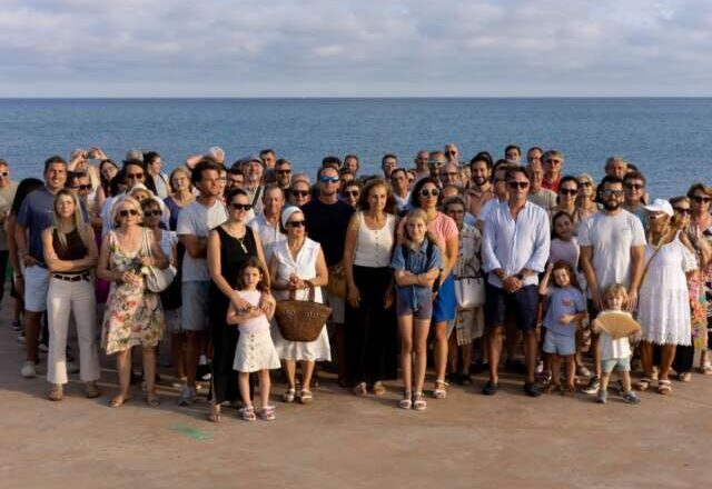 Large group of people posing for a photo on a seaside pier, with the ocean in the background on a sunny day; adults and children of various ages stand together in casual summer clothing.