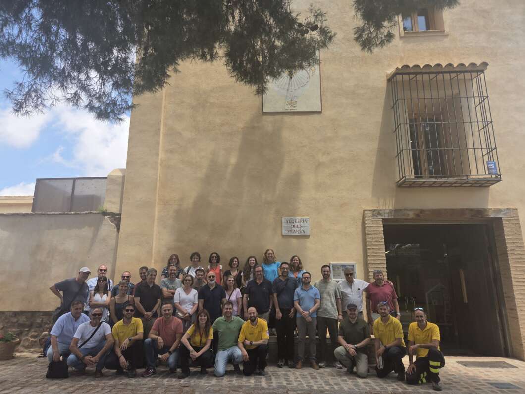 Large group of adults posing for a group photo outside a beige stucco building with a barred window and open doorway on a cobblestone courtyard.