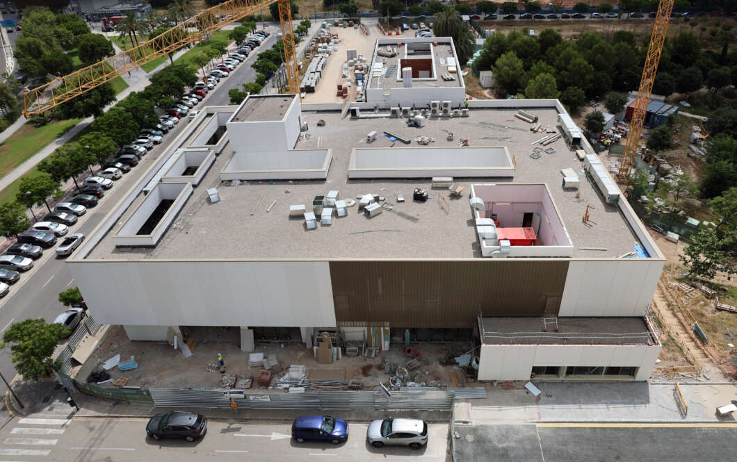 Aerial view of a large construction site on a flat-roofed building, with two yellow cranes, scattered materials, and cars parked along the street.