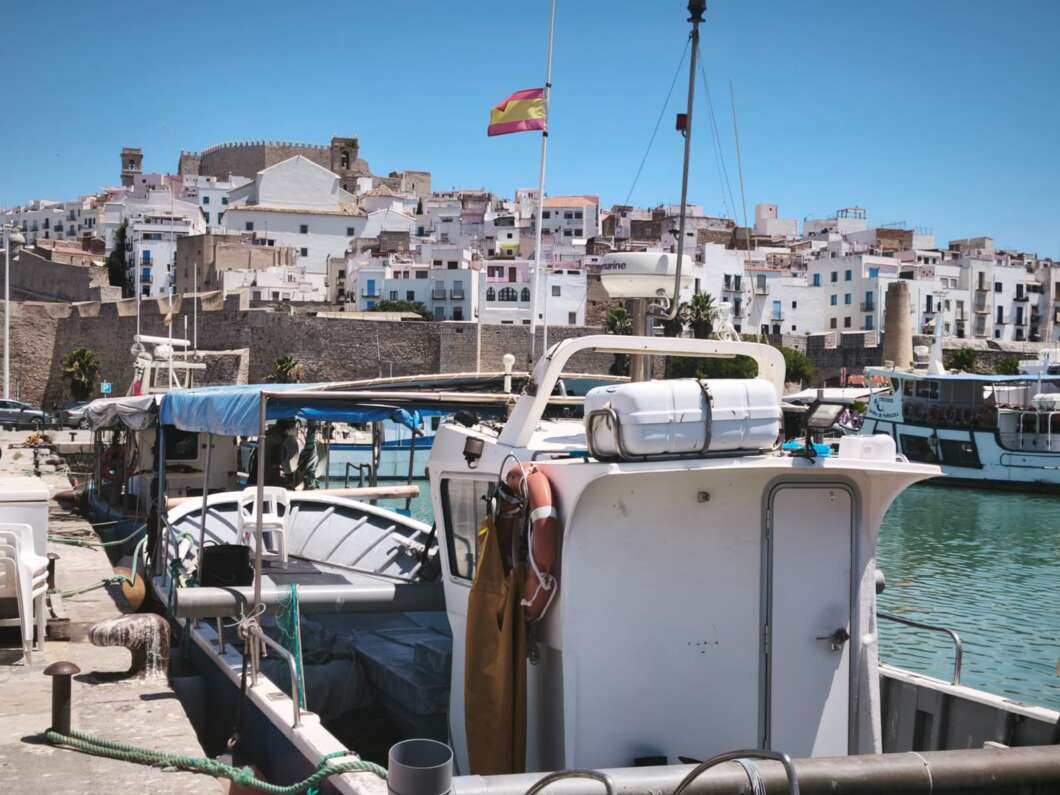 White fishing boat docked at a harbor with pastel houses and a stone wall town in the background under a blue sky, Spanish flag overhead