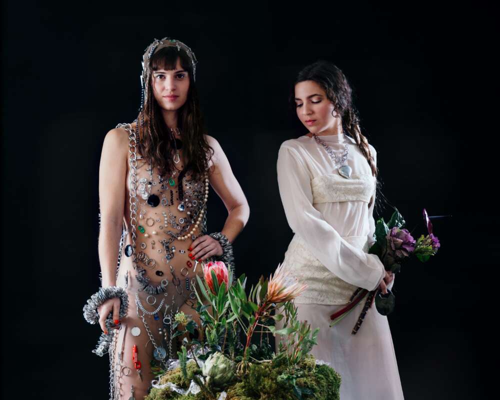Two women pose in a studio wearing avant-garde outfits; one is sheer with jewelry all over, the other in a white dress holding flowers against a dark background.