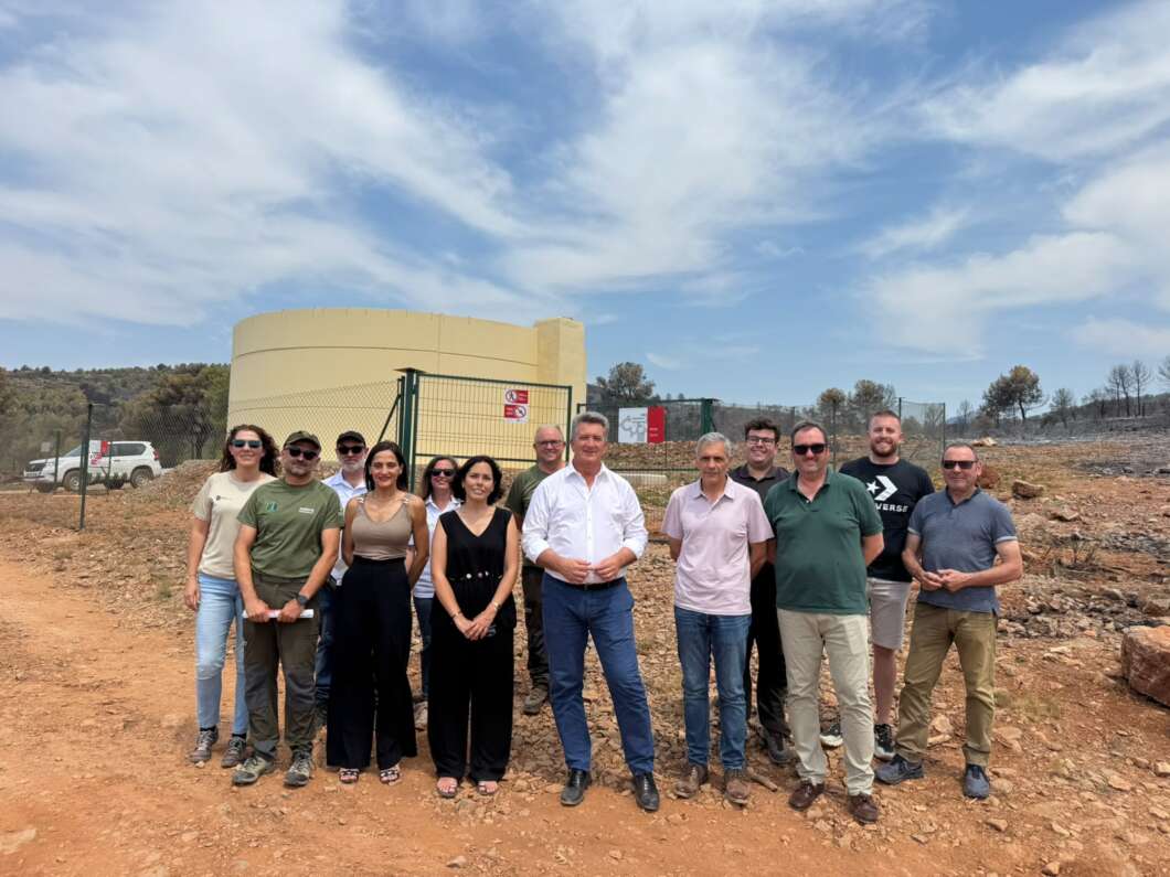 Group of adults posing for a photo at a construction site with a large beige water tank and chain-link fence in the background on a dirt lot.