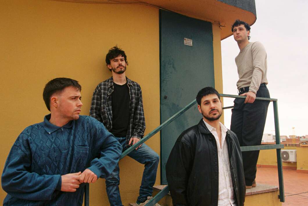 Four men stand on a rooftop stair railing beside a mustard-yellow wall and a blue door, posing casually and looking in different directions.