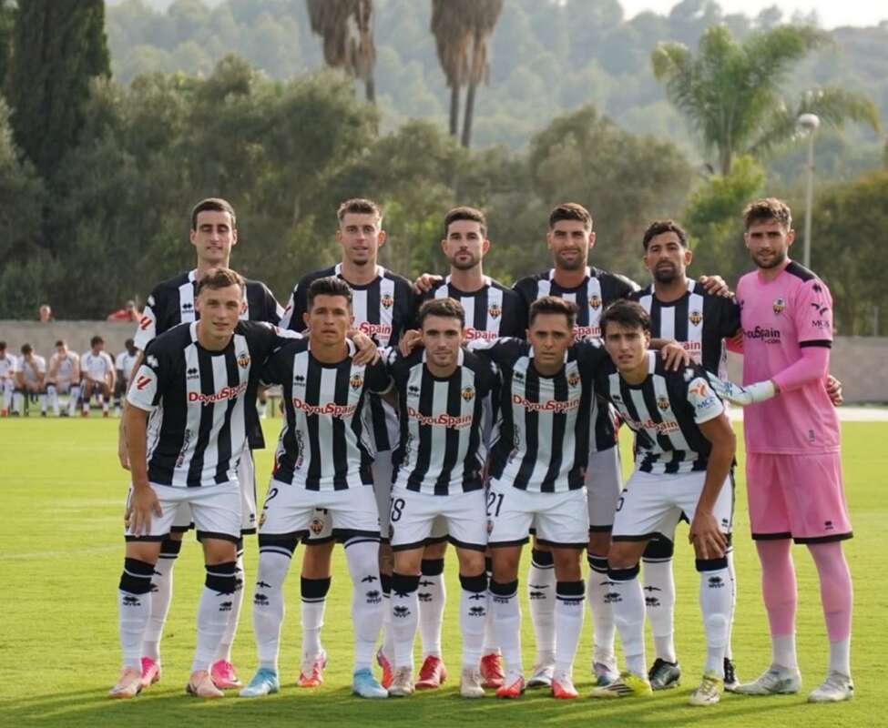 Soccer team in black-and-white striped jerseys posing for a group photo on the field, goalkeeper in pink on the right.