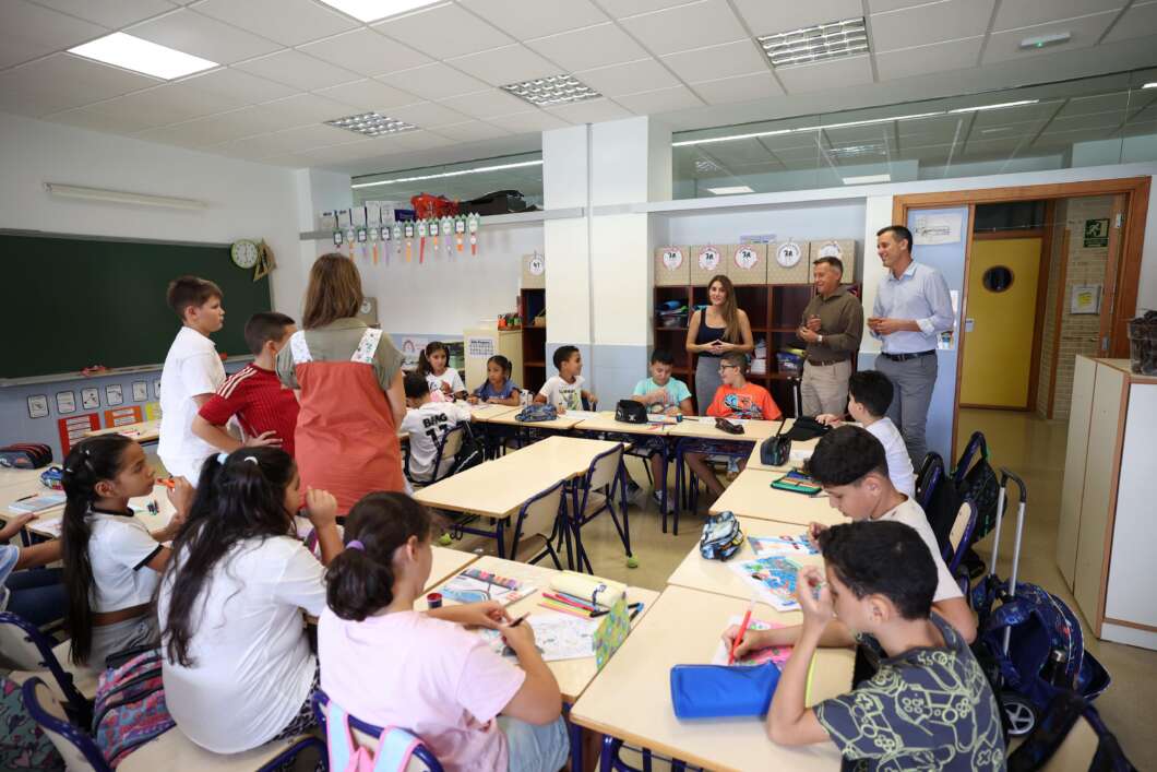 Classroom with students seated around tables as three adults stand at the back observing. Ready-to-color supplies visible on desks.