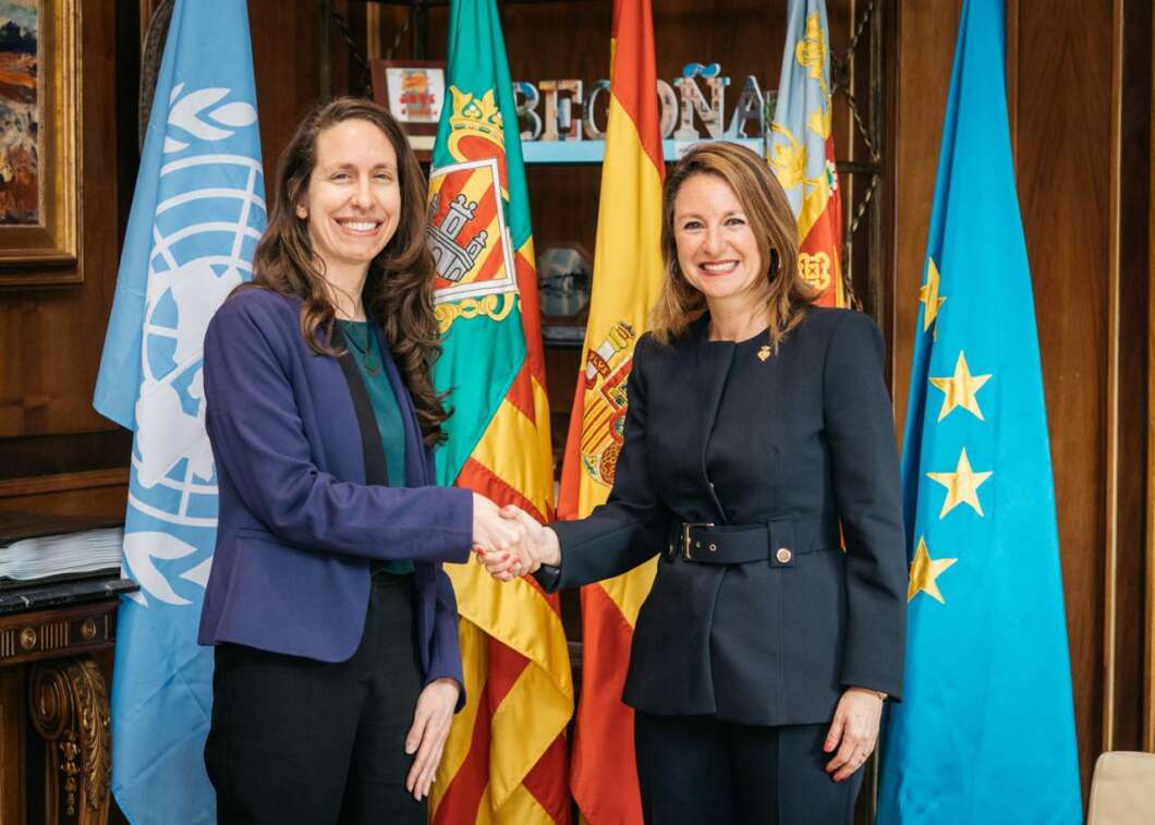 Two women in formal attire shake hands in front of international flags, signaling a diplomatic meeting.