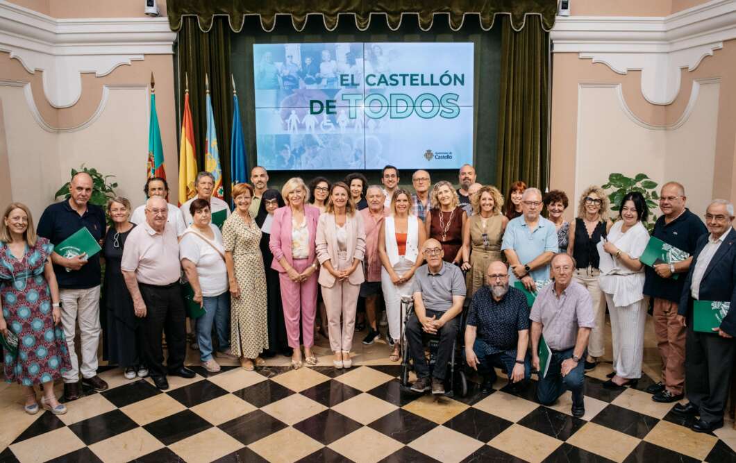 Group of around 30 people posing for a photo in a ceremonial hall with a screen reading 'El Castellón de Todos' behind them; several hold green folders and flags line the back wall.