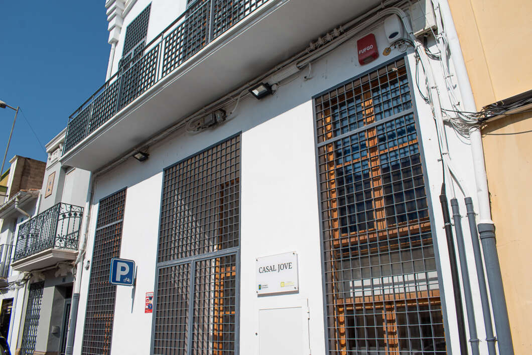 White building with large barred windows and metal balconies on a sunny street; sign reads 'CASAL JOVE' at the entrance.