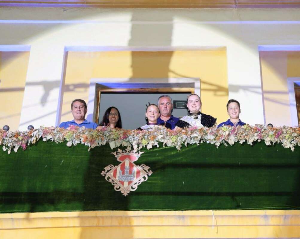 Group of six people leaning over a green floral balcony railing with a crest emblem in front, balcony setting.