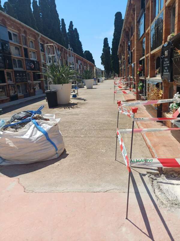 Cemetery path between mausoleums with red-and-white tape along the walkway and a large white bag of debris on the left under a clear blue sky.