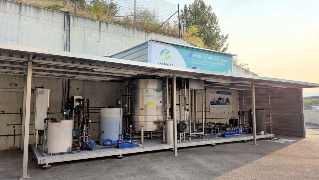 Industrial facility with stainless steel tanks, pipes and pumps on an elevated platform under a shed.