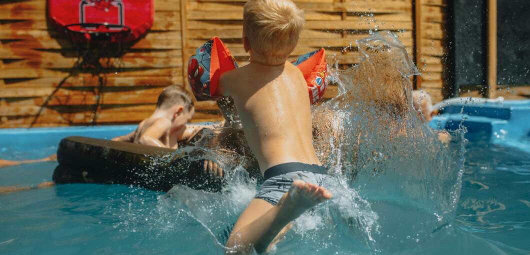 Children playing in a sunny backyard pool; a boy with inflatable armbands jumps, splashing water.