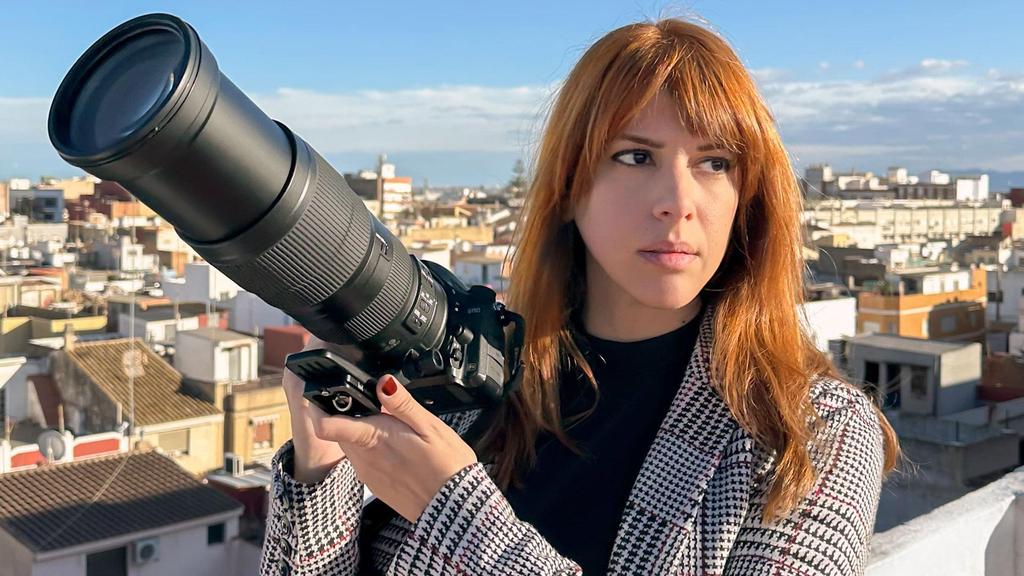 Woman with red hair holding a large telephoto camera on a rooftop overlooking a cityscape at dusk/dawn light