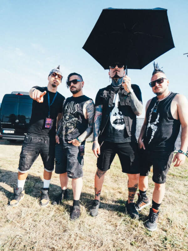 Four tattooed men in black rock attire posing outdoors at a sunny event, one holding a large black umbrella.