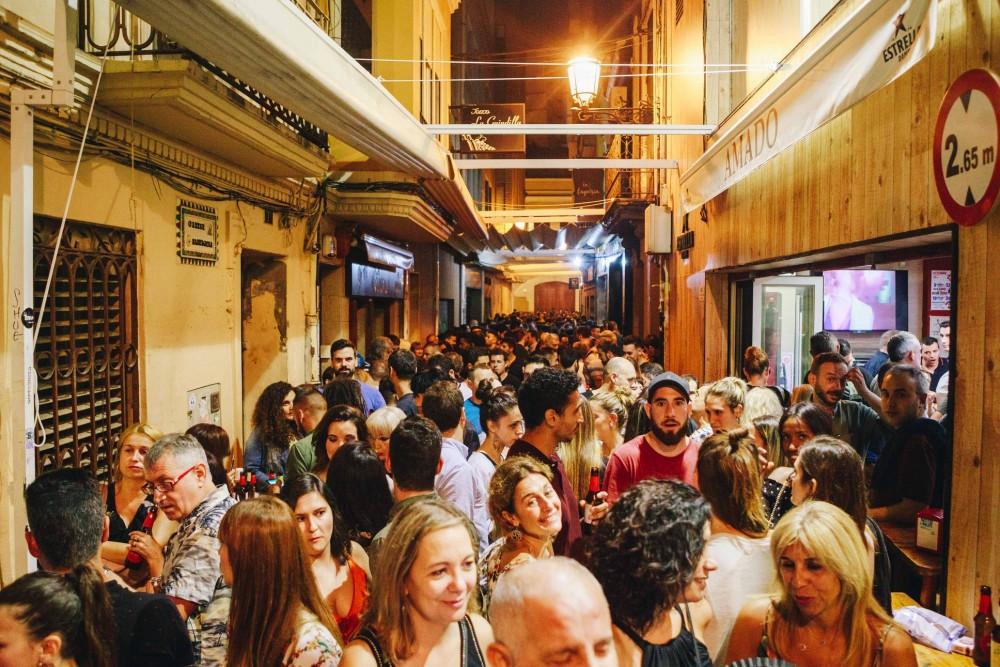 Crowded nighttime street alley filled with people socializing and holding drinks under warm lights between shopfronts and bars.