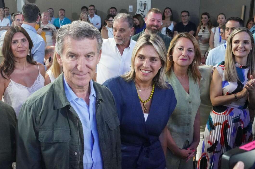Group of smiling adults at an outdoor event, with a man in a green jacket and a woman in a blue dress at the front center.