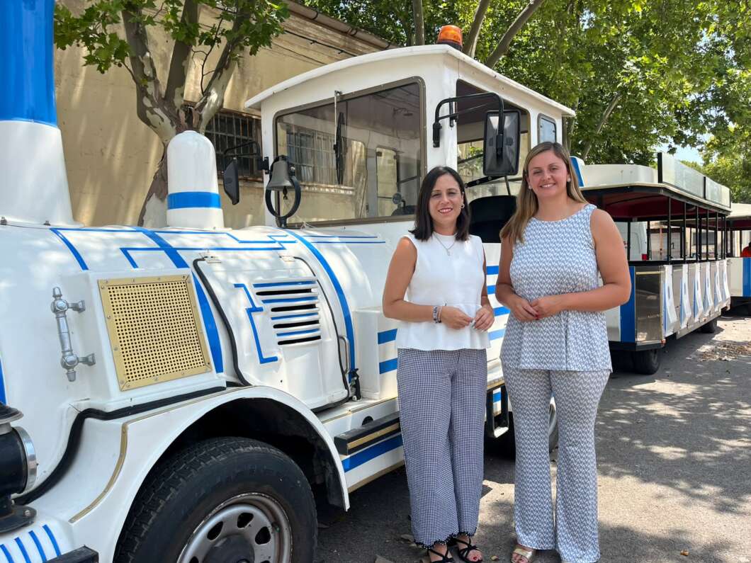 Two women stand beside a white and blue tourist train on a sunny street, smiling, with the train’s driver cabin and connected passenger cars in the background.