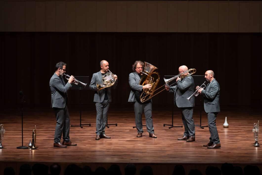 Five male musicians in gray suits perform a brass quintet on a wooden stage, each playing a brass instrument (trumpet, horn, tuba, trombone, trumpet).