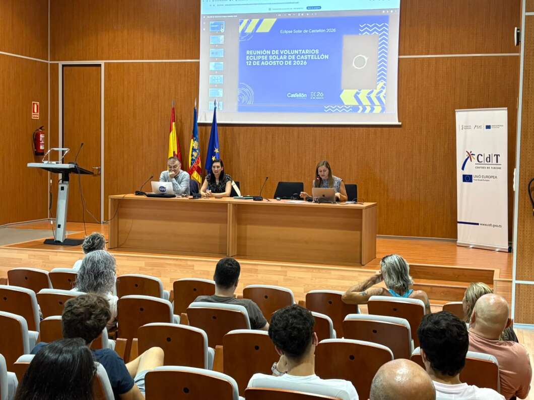 Panel of three speakers seated at a long wooden desk on a stage, presenting to an audience with a projected slide and flags in the background.