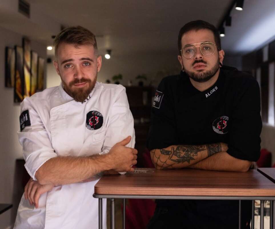 Two chefs lean on a wooden counter in a cafe, one in a white jacket and the other in a black shirt, looking at the camera.