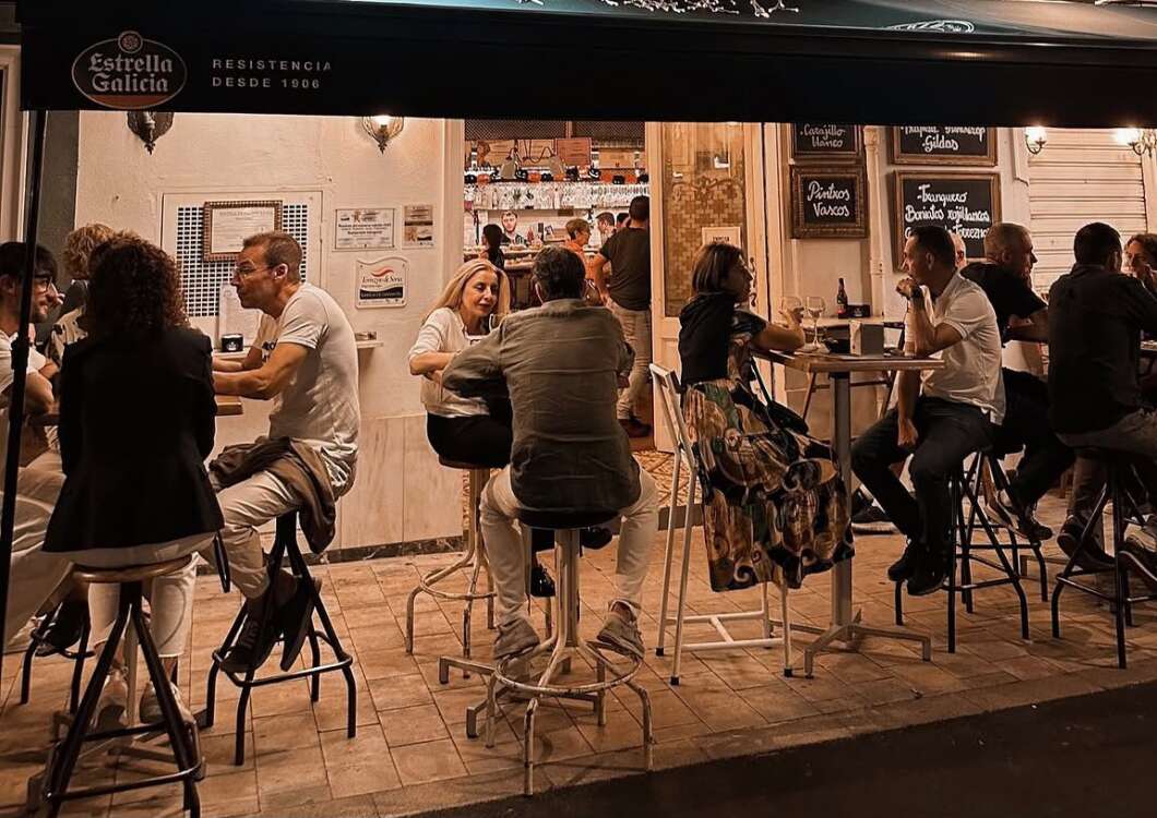 Outdoor bar scene at night with groups of people sitting on high stools, talking and drinking; Estrella Galicia branding visible above.