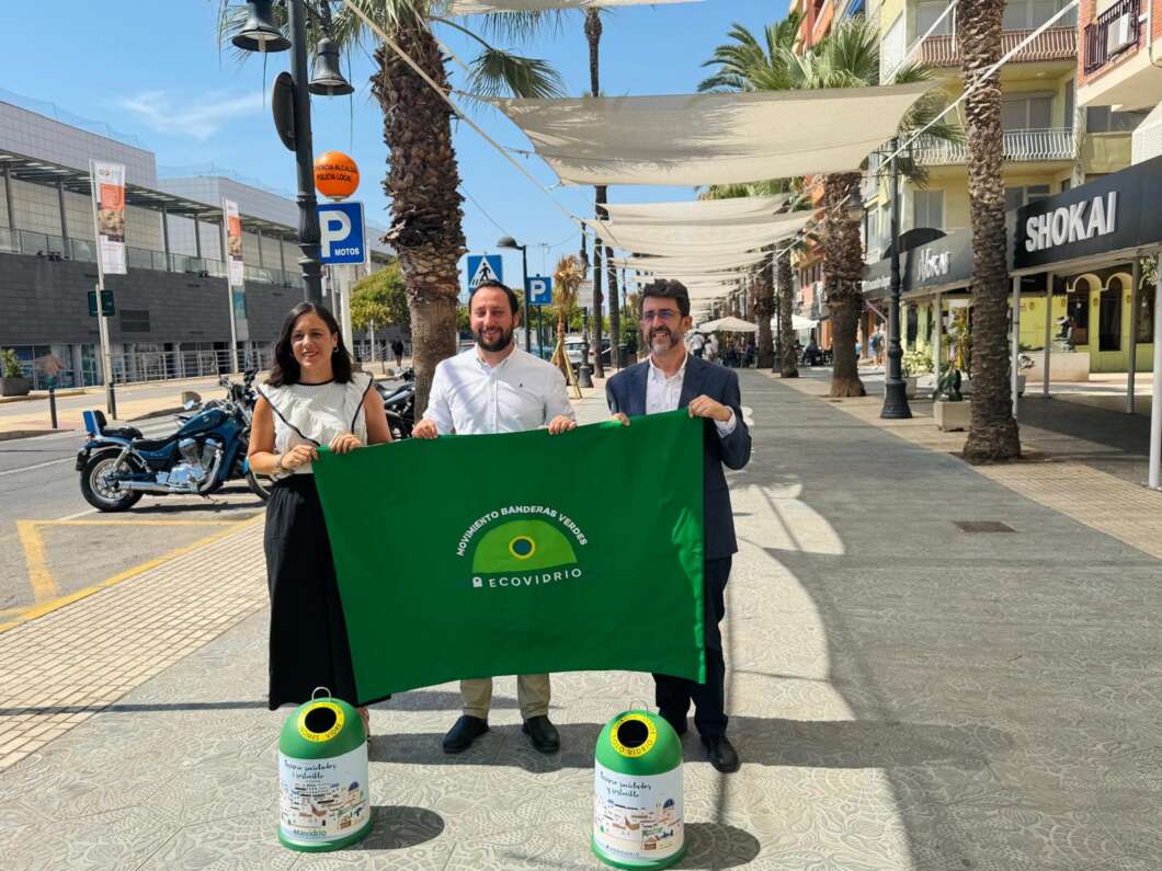 Three people stand on a sunny pedestrian street, holding a green banner that reads 'Movimiento Banderas Verdes – Ecovidrio,' with two green cylindrical recycling bins at their feet.
