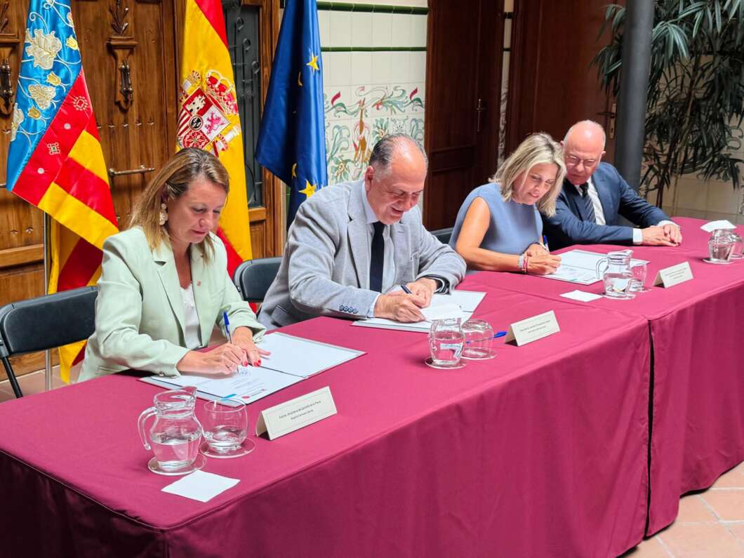 Four professionals sign documents at a formal signing ceremony, with national flags in the background and water pitchers on a maroon tablecloth.