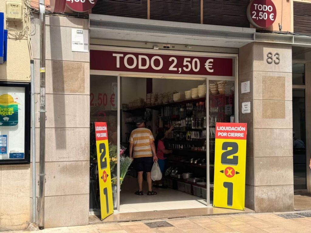 Front of a small shop with a maroon sign reading 'TODO 2,50 €' and tall yellow sale signs outside that say 'LIQUIDACIÓN POR CIERRE' and '2 1'. Inside, a person browses shelves of items.