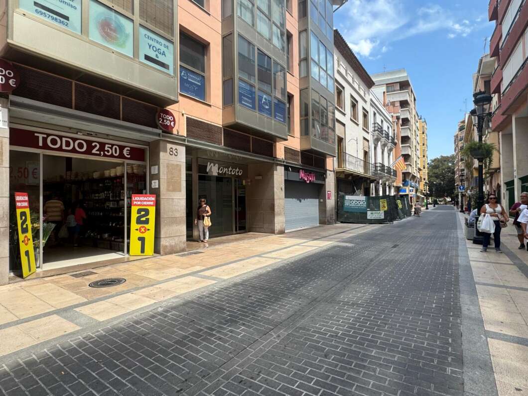 City street with shops on both sides and a few pedestrians walking along a sunny pedestrian avenue; store signs visible, including 'TODO 2,50 €'.
