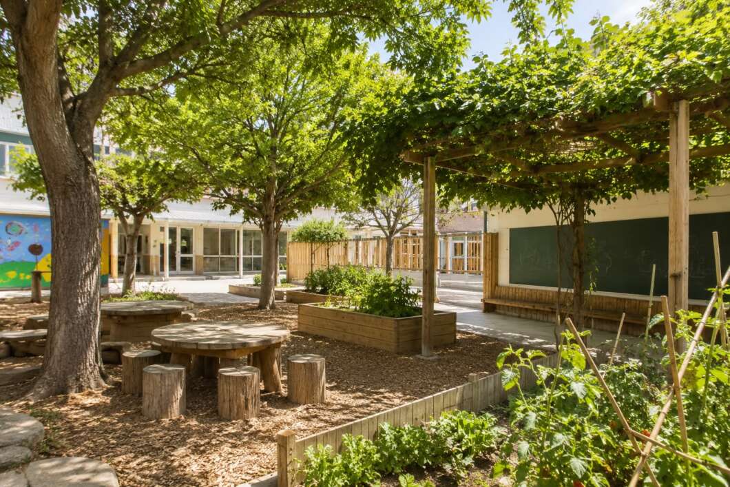School courtyard with shaded wooden tables and tree-stump stools, raised garden beds, and a vine-covered pergola.