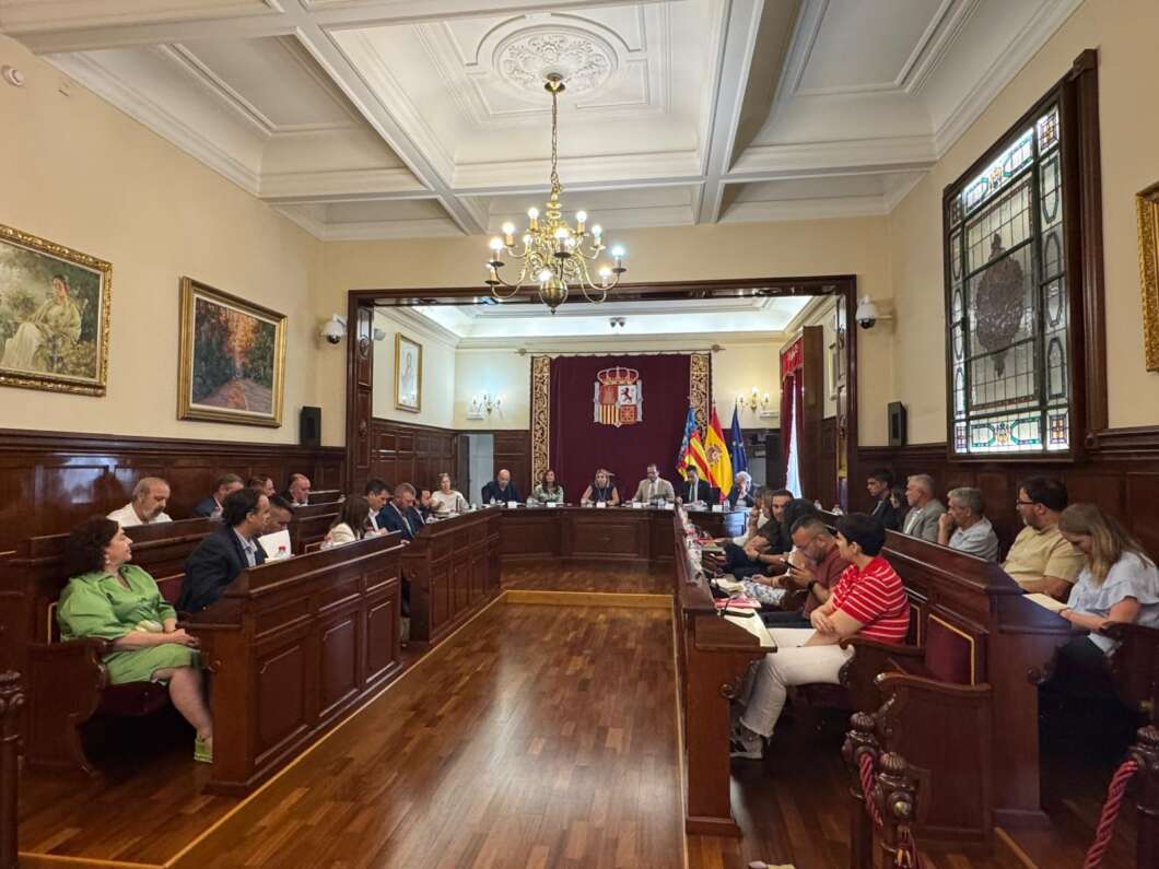 Formal council chamber with attendees around a U-shaped wooden table, a chandelier overhead, and national/regional flags in the background.