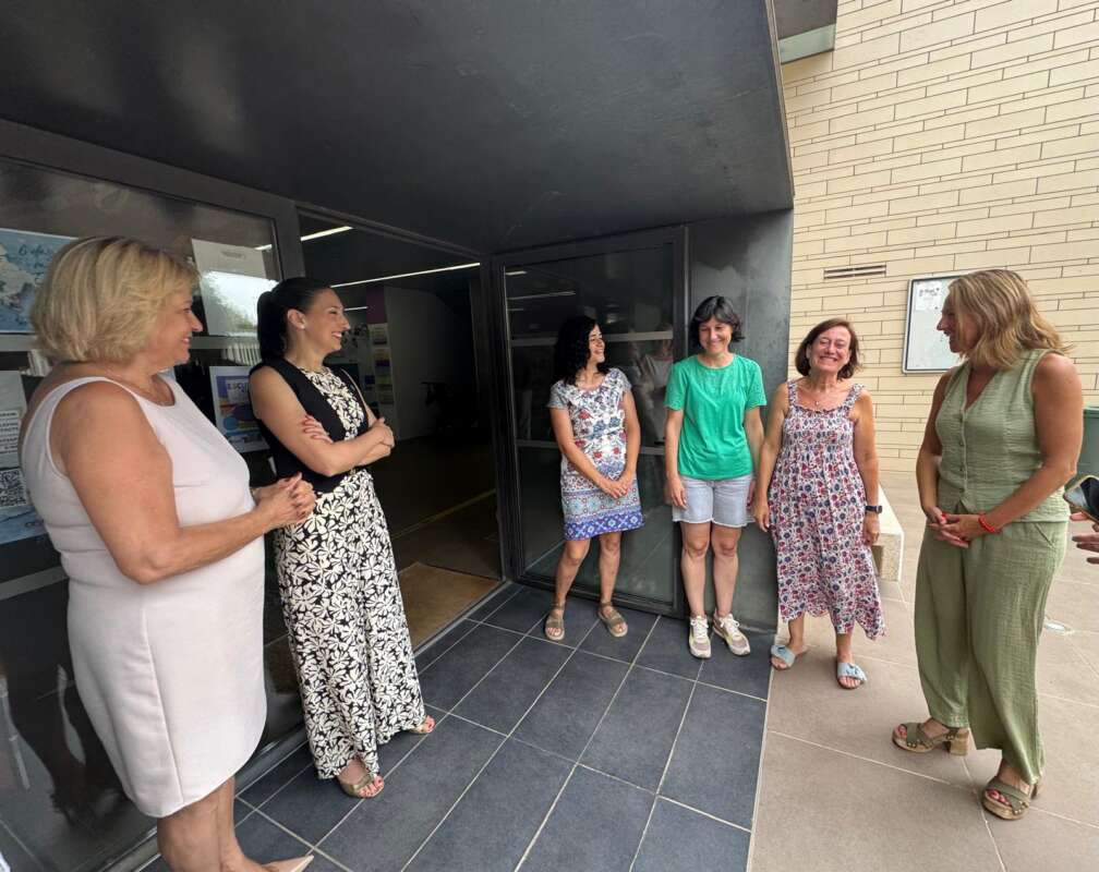 Six women stand in a semicircle outside a glass entrance, chatting and smiling.