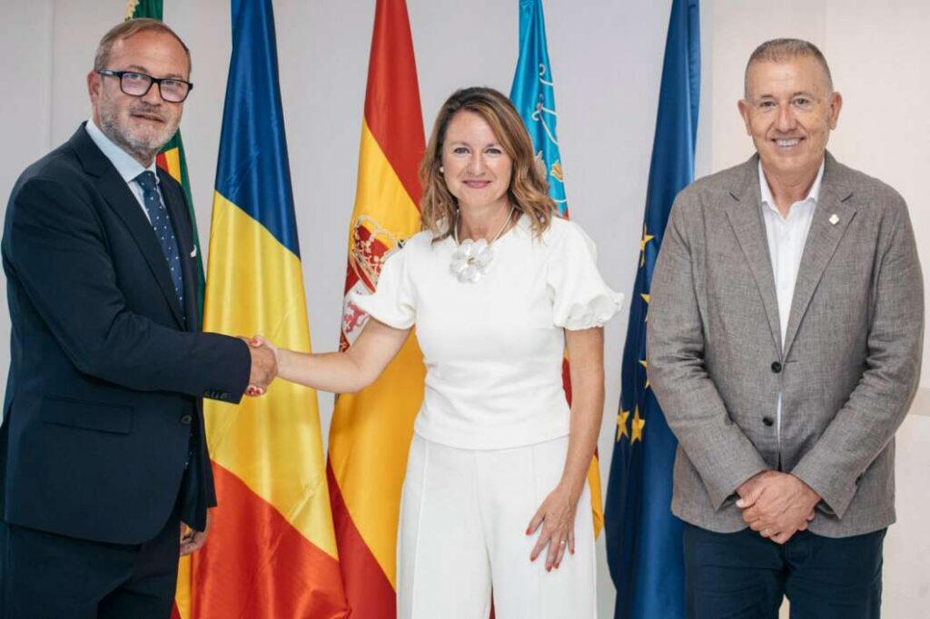 Three professionals stand in front of flags; the man on the left and the woman in the center shake hands, while the man on the right smiles.