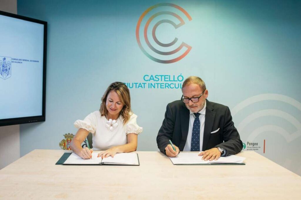Two professionals sign documents at a table during a formal signing ceremony, with a blue backdrop and logos behind them.