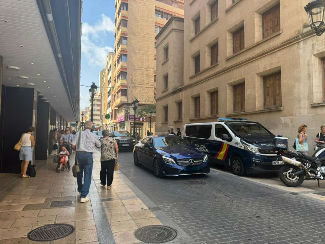 Street scene with a blue Mercedes patrol car and a Policía Nacional van parked along a cobblestone street; pedestrians walk on the sidewalk.