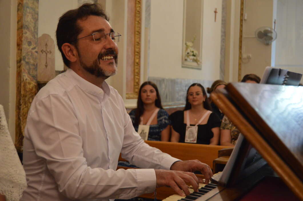 Pianist in a white shirt smiling while playing a piano in a church, with two women seated in the background.