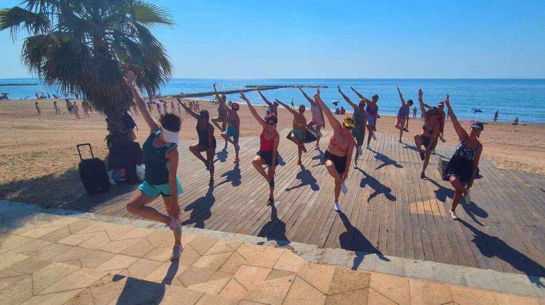 Group of people doing yoga on a wooden deck by the beach, arms raised in tree pose under a palm tree with the sea in the background.