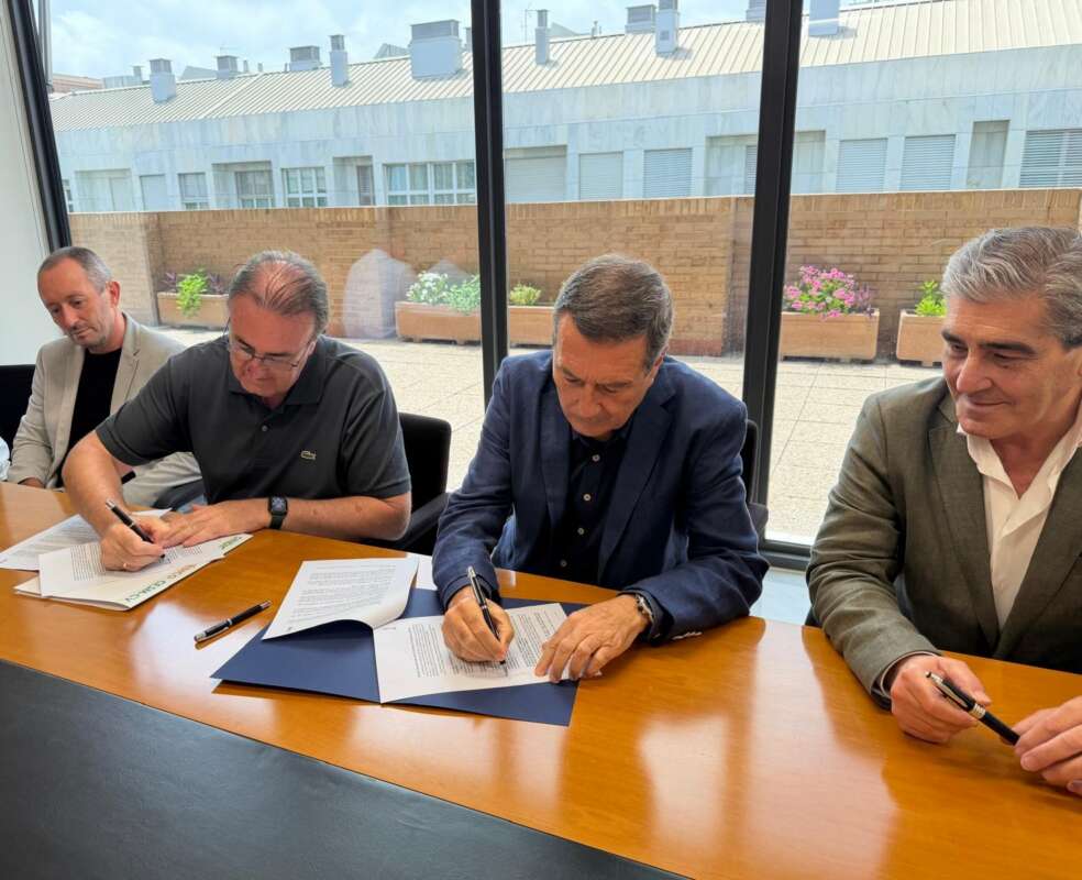 Four men sit at a conference table signing documents during a meeting in a bright room with large windows behind them.