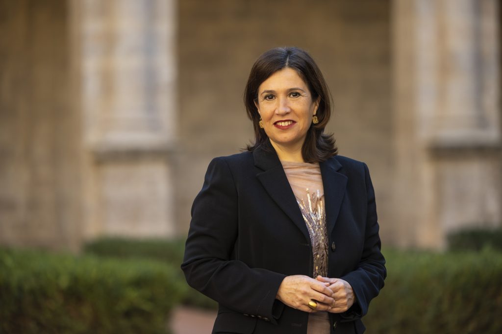 Professional woman in a dark blazer stands outdoors, smiling, with blurred stone columns in the background and hands loosely clasped.