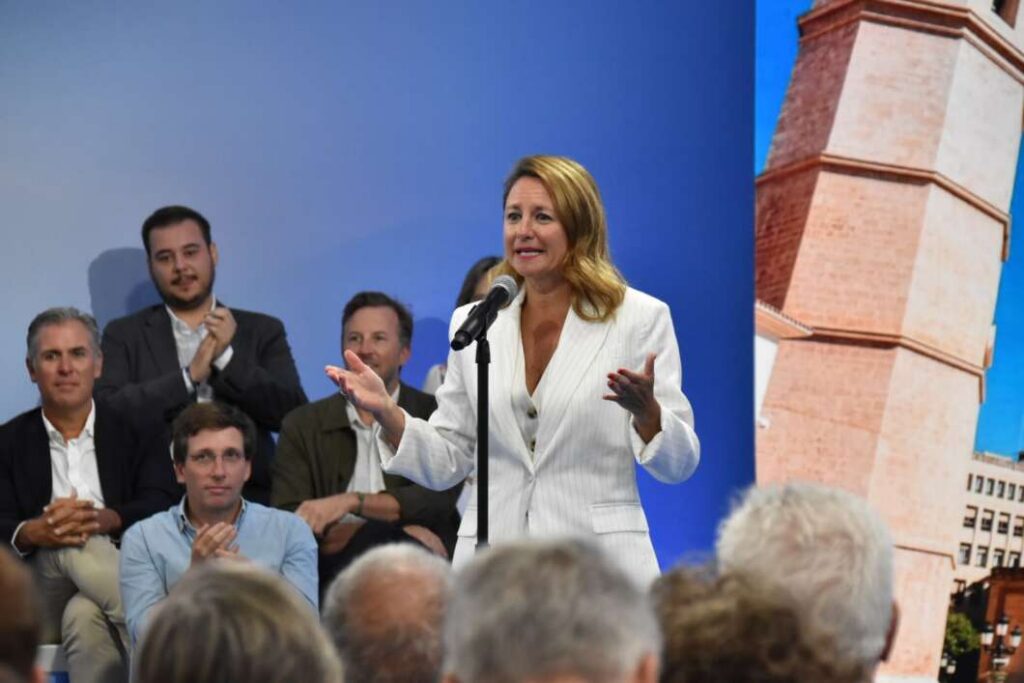 Female speaker in a white blazer addresses the audience at a microphone on stage, gesturing with hands.