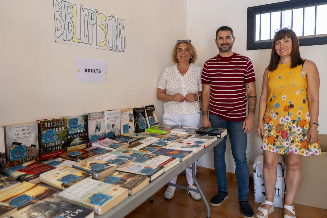 Three adults stand behind a long table filled with books at a community book sale, with an 'ADULTS' sign on the wall behind them.