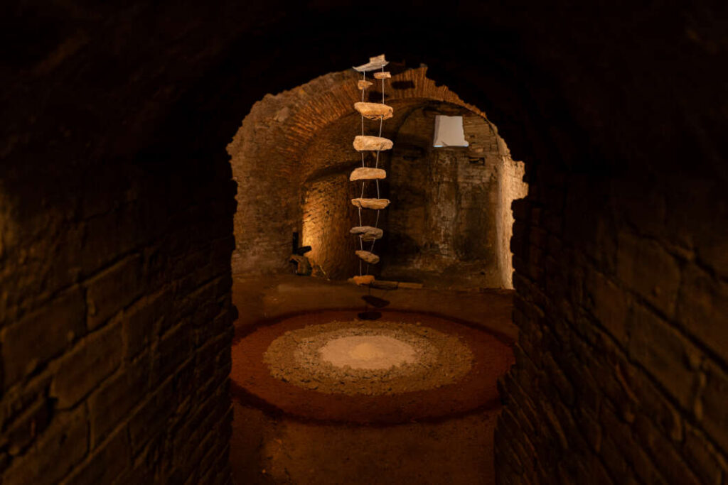 Underground brick tunnel with a hanging spiral ladder and a circular sand ring on the floor, dimly lit by warm light in the background.