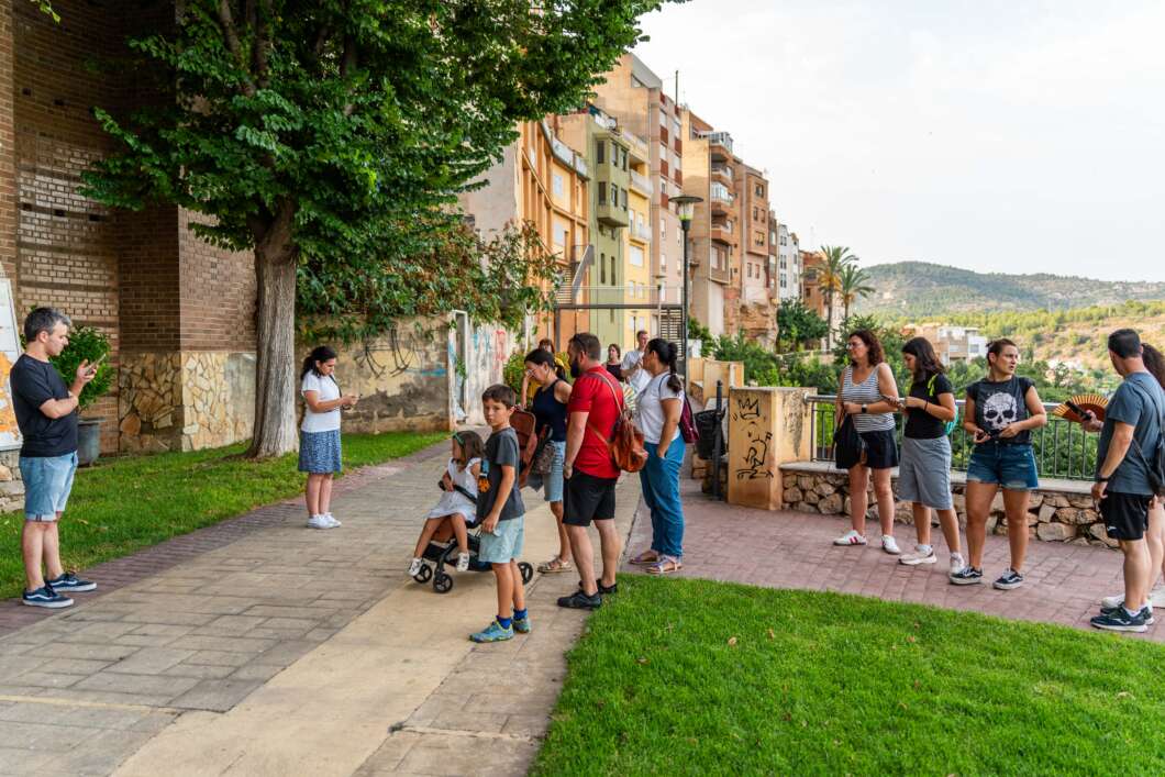 People stand in a line along a paved park path beside a green lawn, with apartment buildings in the background on a sunny day.