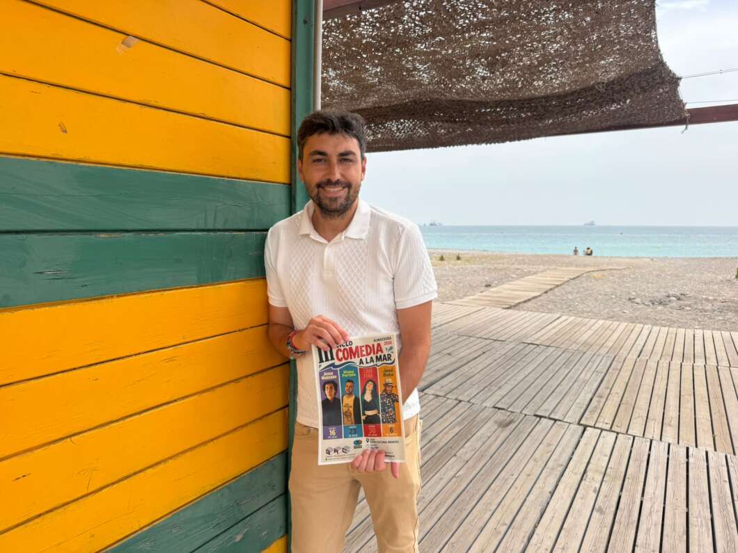 Man in a white polo and khaki shorts stands beside a yellow-green beach shack, holding a poster for 'Comedia a la Mar' with performer photos at the bottom. The beach and ocean are in the background.