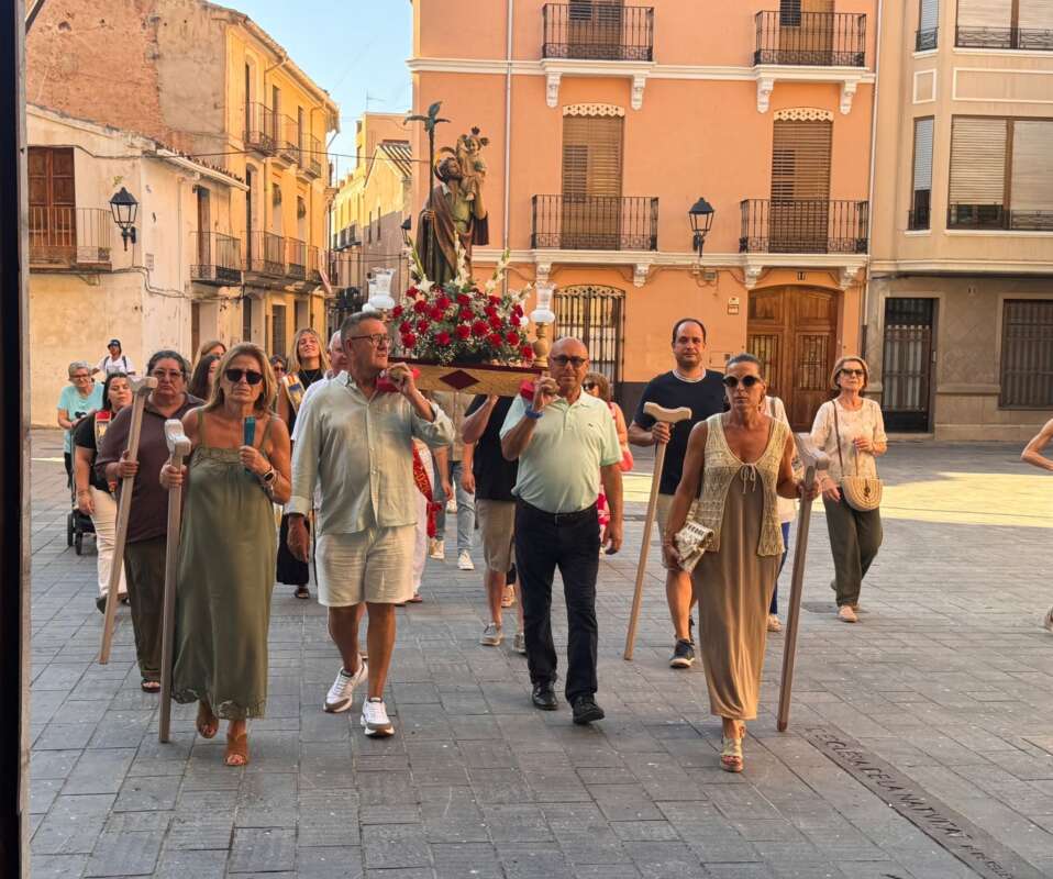 Religious procession: people carry a flower-decked statue on a platform through a sunlit street.