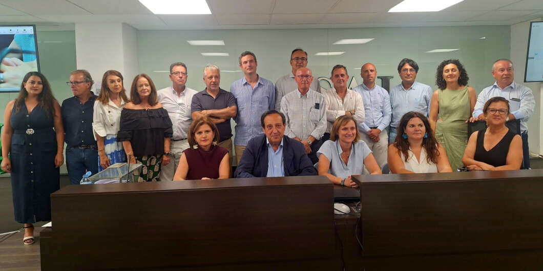 Group of colleagues in an office, some seated at a long desk and others standing behind, smiling for a team photo in a conference room.