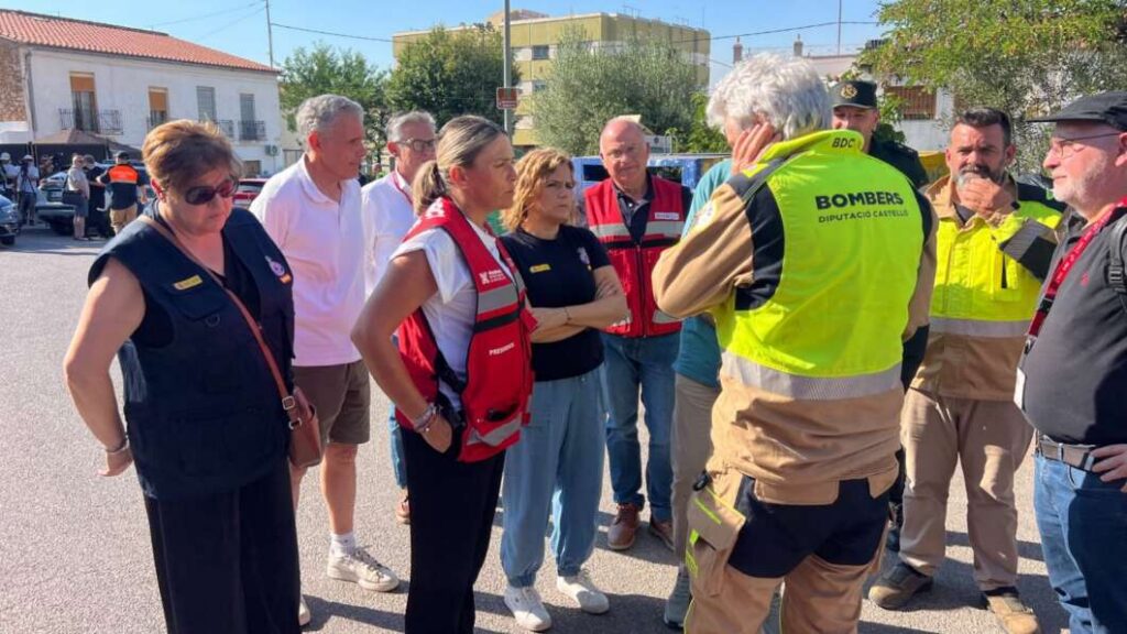 Group of emergency responders and volunteers gathered in a circle on a sunny street, listening to a briefing; one person wears a bright yellow safety vest labeled 'BOMBERS'.