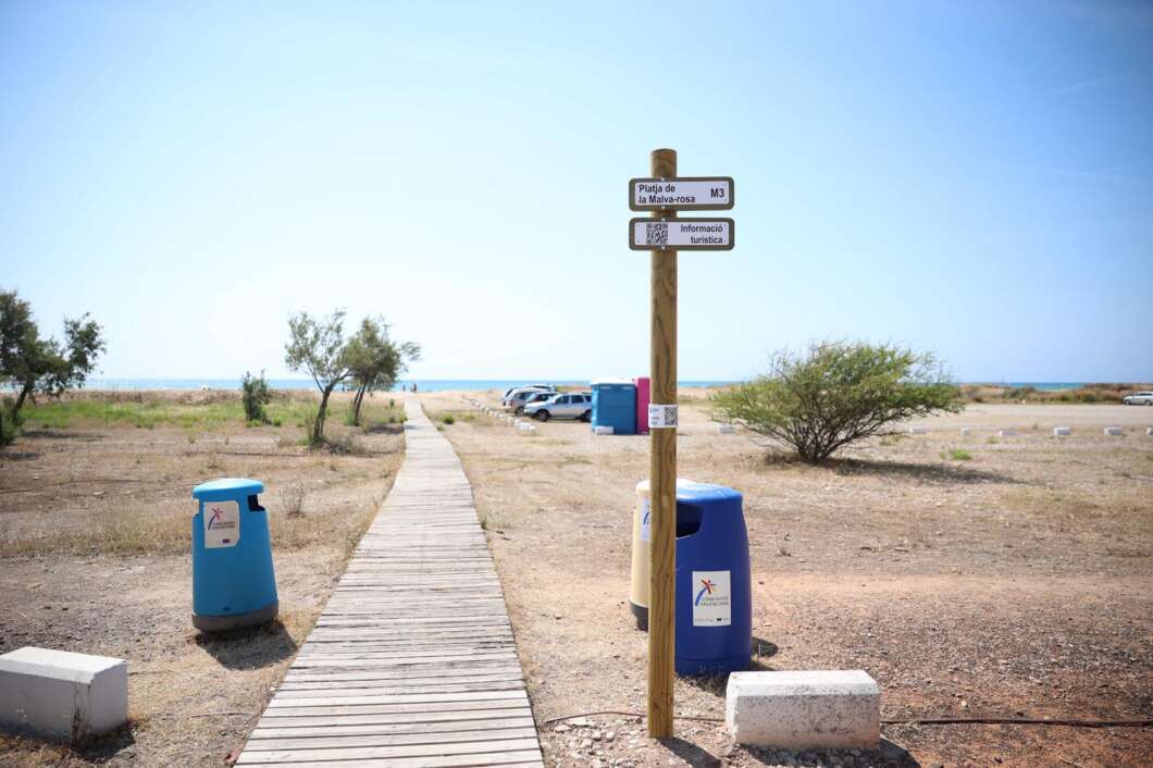 Wooden boardwalk leads to a sunny beach with a signpost and blue trash/recycling bins nearby, flat coastal landscape in the distance.