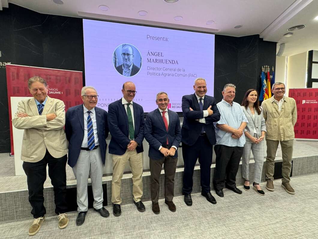 Group of nine adults in business attire standing on a stage for a presentation, with a large screen behind showing a speaker's photo and name Ángel Marhuenda.
